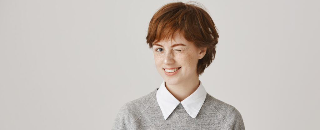 if you know what i mean. studio shot of positive attractive redhead woman with freckles winking as if being on same line with friend, agreeing with idea, standing over gray background